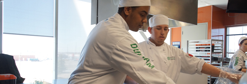 Two male students in a professional kitchen wearing chef hats and coats